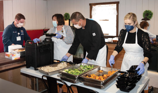 Four people wearing face masks and gloves stand at a table with food trays. They are preparing meals in black containers.