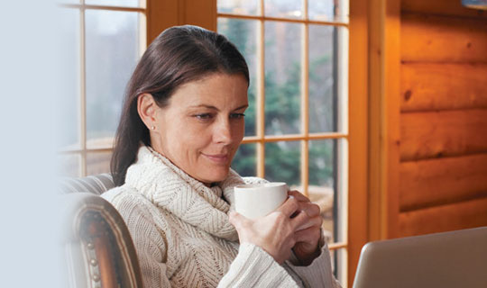 woman sitting on a comfy chair sipping from a mug while looking at her laptop