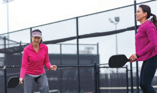 2 women in bright pink shirts playing pickleball on an outdoor court