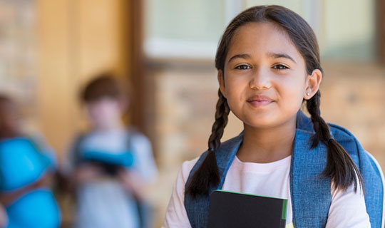 young girl wearing a backpack and holding a notebook at school