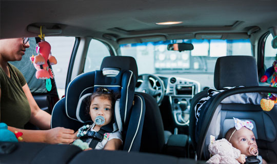 two young children in car seats in the back of a car - one car seat is being checked for safety