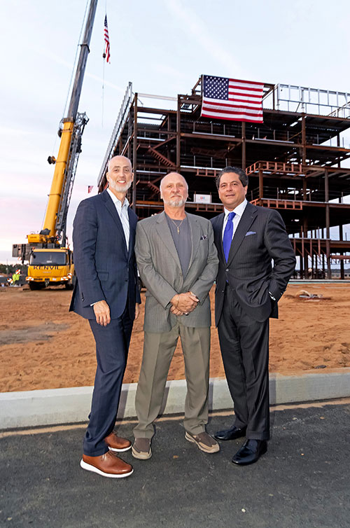 Eric Carney, Tommy Mears, and Mark E. Manigan standing in front of the building as the final beam is placed at the Specialty and Cancer Care Pavilion at the Vogel Medical Campus in Tinton Falls